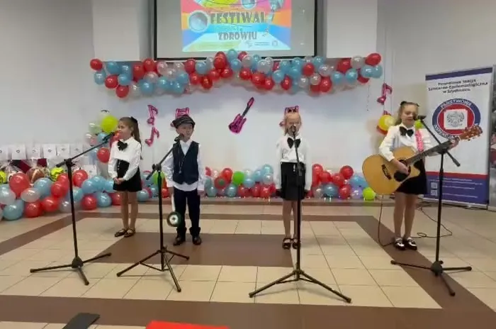 Czwórka uczniów w eleganckich strojach, jedna dziewczynka gra na gitarze